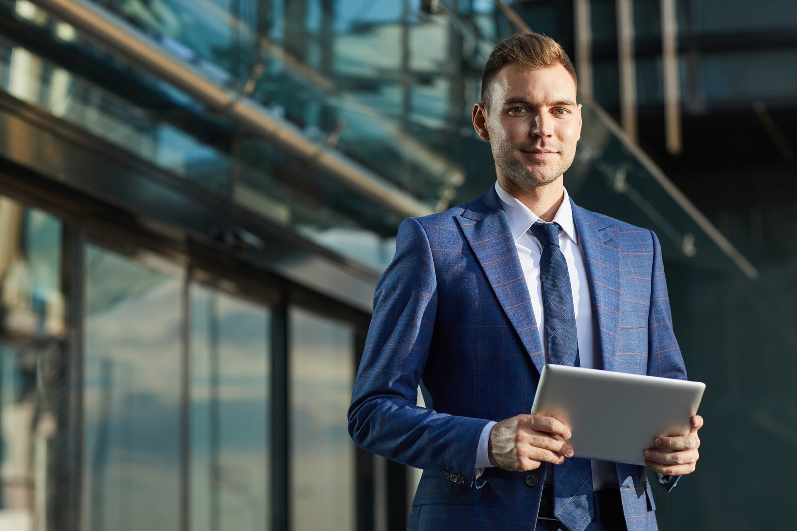 Portrait of young businessman in formal suit looking at camera while using digital tablet in the city